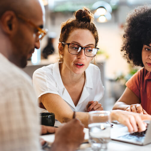 Three diverse small business partners collaborate and discuss ideas around a table with a laptop and glasses of water.