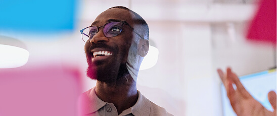 A smiling man with glasses and a beard looks upward in a bright, modern workspace with pink and blue accents.