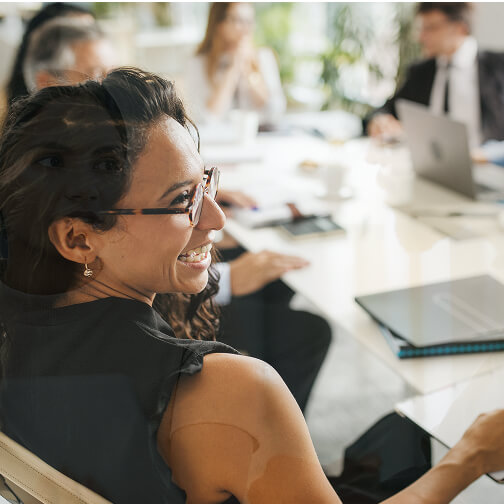 A smiling woman with glasses sits at a conference table with colleagues in a professional meeting setting.