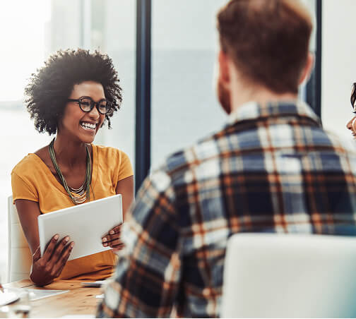 A smiling woman with glasses holding a tablet discusses with two clients in a professional office setting.