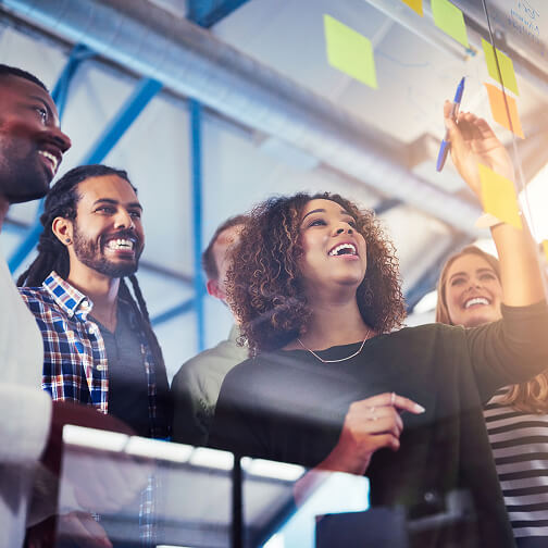 A diverse group of people happily collaborating and writing on a glass wall with sticky notes in a professional setting.