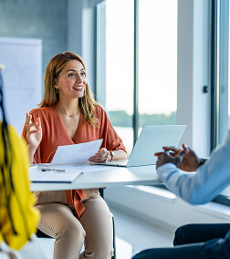 Diaconia Who We Serve PreFooter A woman with long red hair smiling and speaking during a meeting in a bright office with large windows.