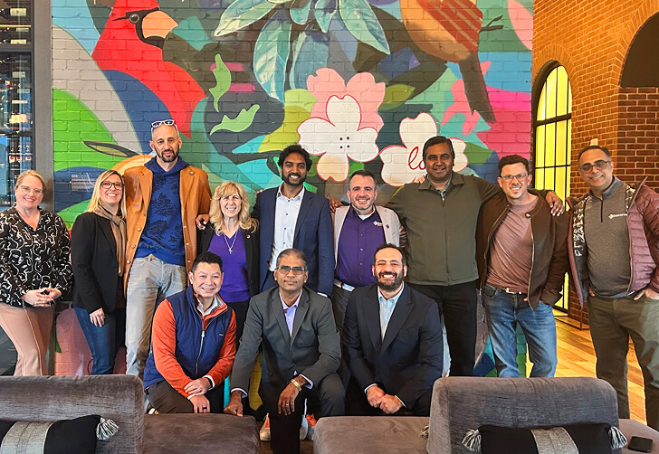 A diverse group of ten people poses together in front of a colorful mural with flowers and birds in an indoor setting.