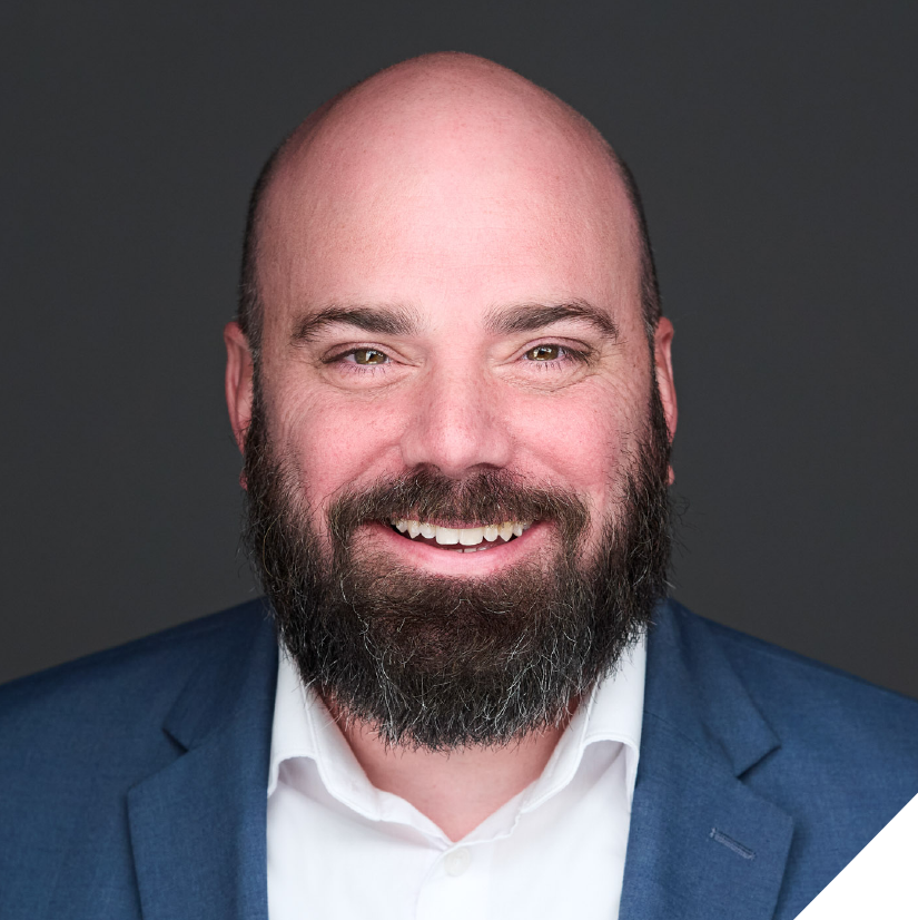 A smiling man with a beard and bald head wearing a navy blazer and white shirt against a dark background.