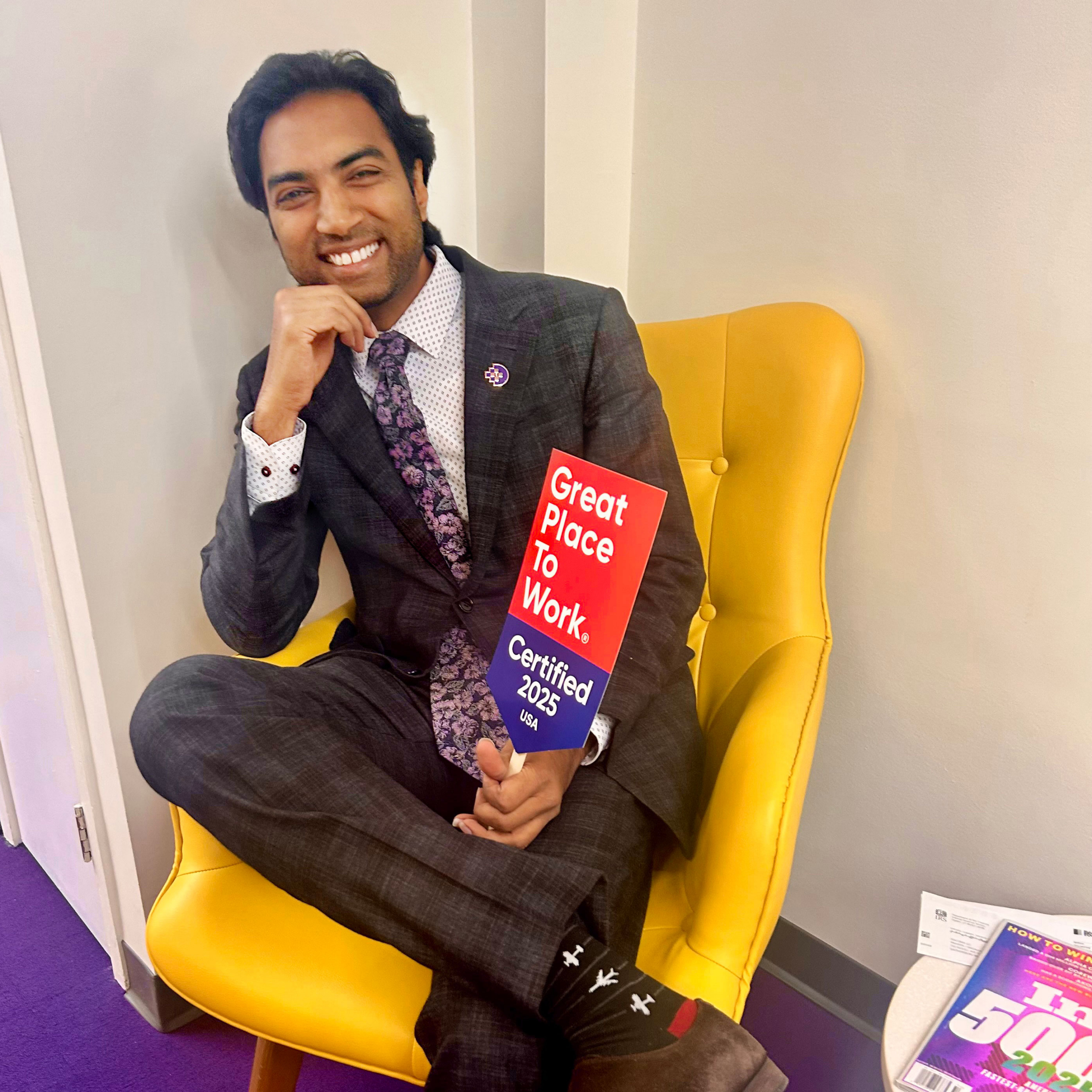 A man in a suit with a patterned tie sits on a yellow chair, smiling and holding a "Great Place to Work" certificate.