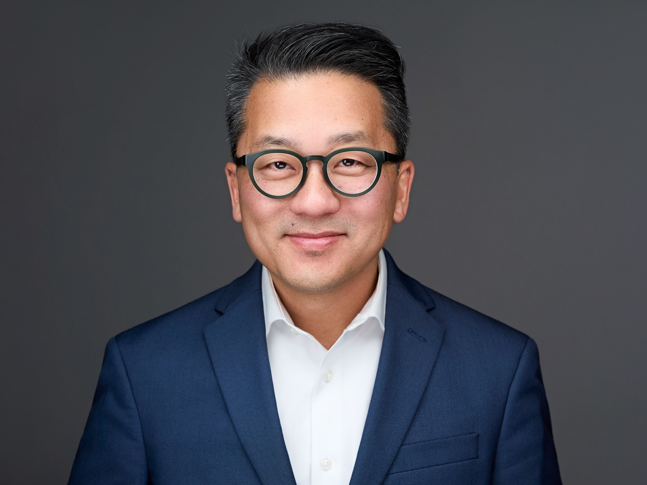 A man with short black hair, glasses, and a friendly smile wearing a navy blue suit and white shirt against a gray