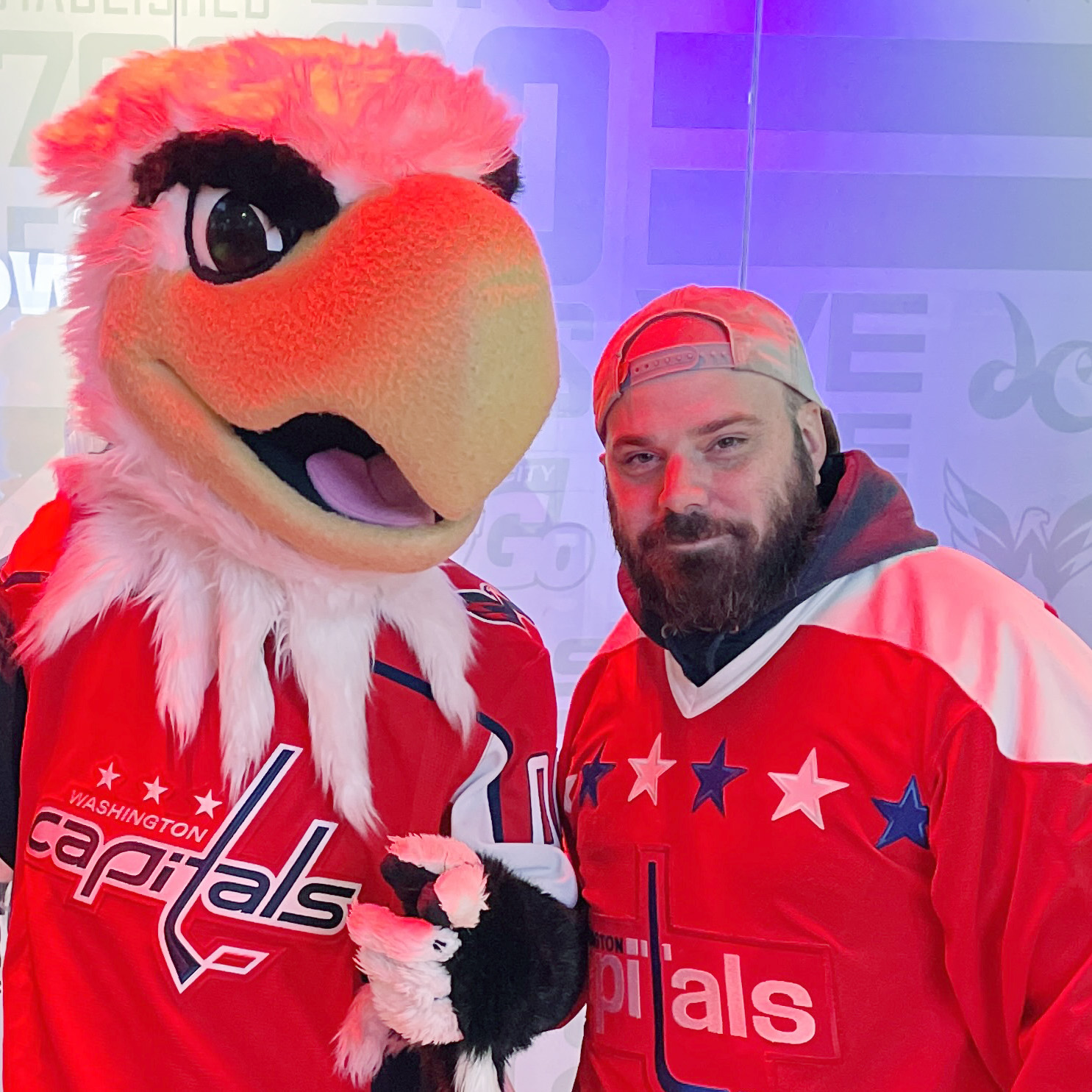 A person in a Washington Capitals hockey jersey stands next to a mascot with a large, colorful bird head and a red cap.