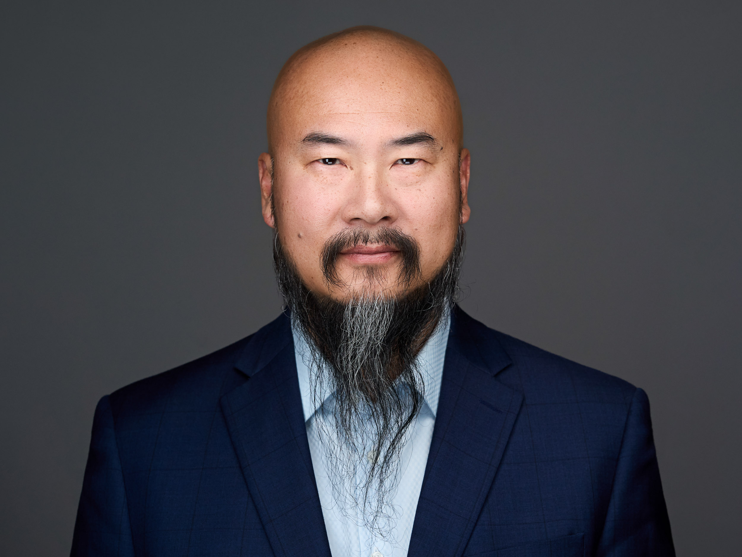 A bald man with a long beard, wearing a dark blue suit and light blue shirt, looks confidently at the camera against a gray